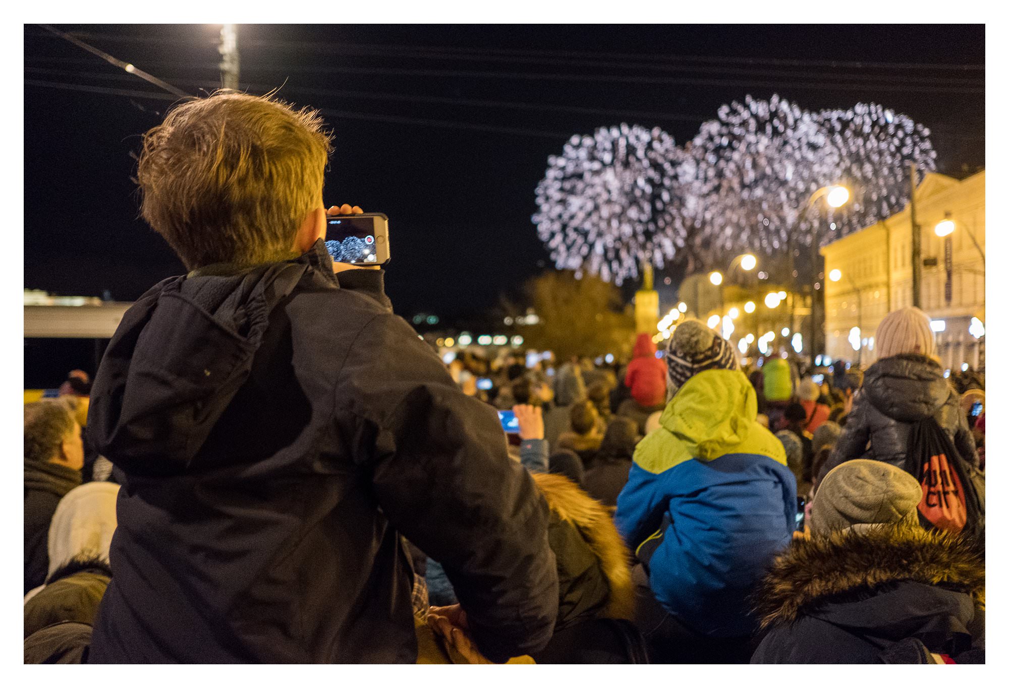 little boy watching New Years eve fireworks on brother's shoulders in Prague while filming on his iPhone
