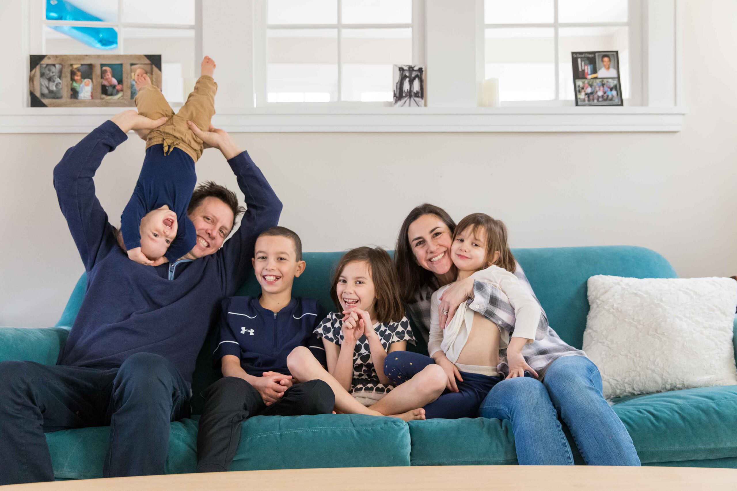 family sitting together on couch, laughing while dad holds baby upside down