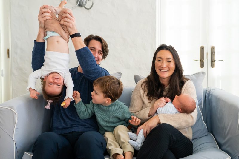 family with two little kids sitting on a couch with a newborn.  dad is holding a toddler upside down