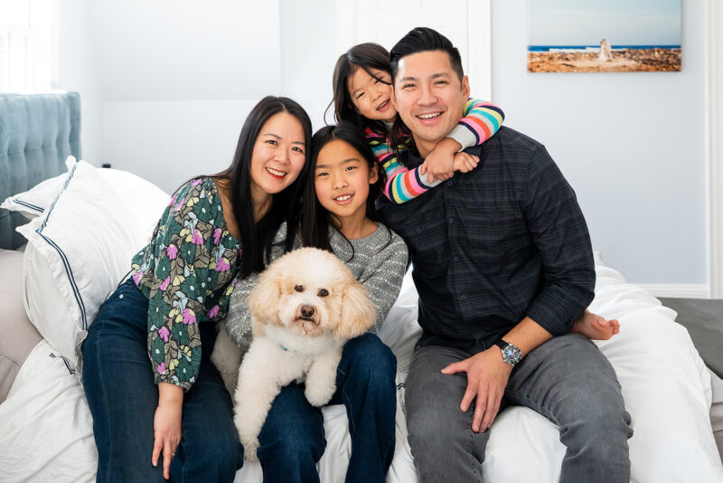A family with two girls sitting on a bed with a white dog smiling at the camera for a family portrait by photographer Jaye McLauglin.