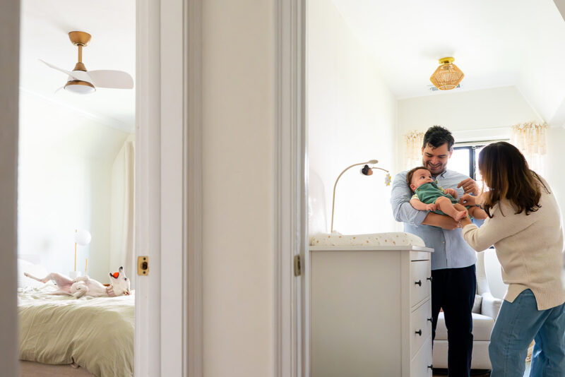 Mom and dad changing a baby with a white dog on the bed who has a ball during a family photo session by Jaye McLaughlin.
