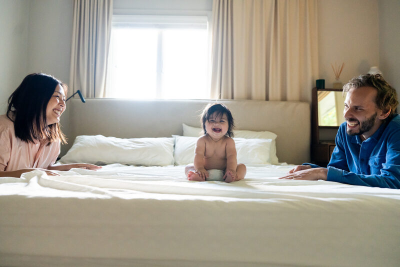 Mom and dad smiling and looking at a baby on the bed during a relaxed in home family photoshoot in Westchester.
