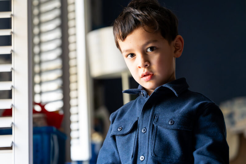 Young boy wearing a blue shirt standing near a window during an in home family photoshoot.